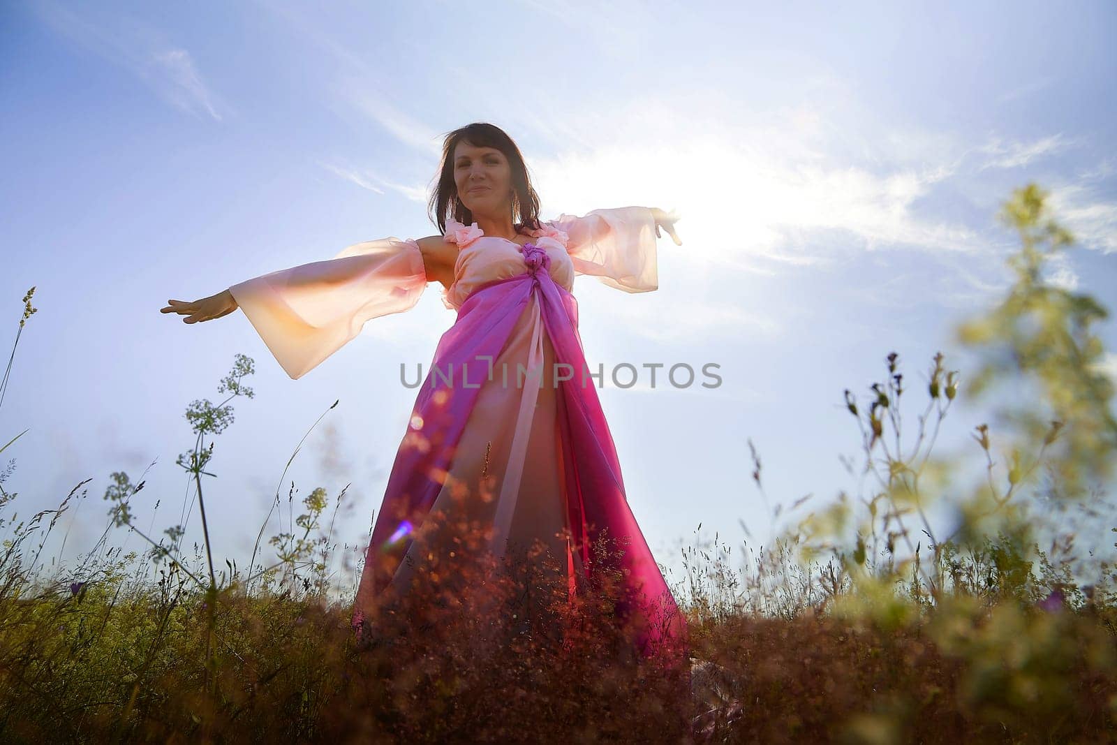 Beautiful girl in lush pink ball gown in green field during blooming of flowers and blue sky on background. Model posing on nature landscape as princess from fary tale