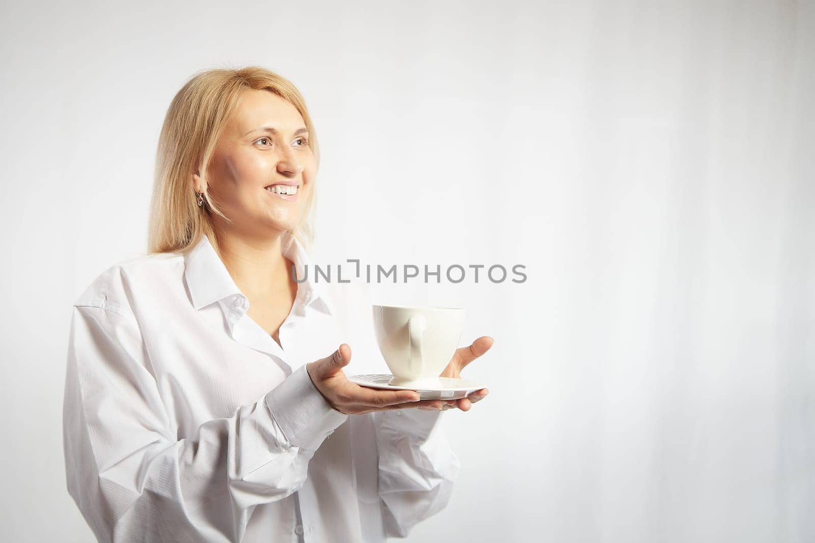 Portrait of pretty blonde smiling woman posing with cup of tea or coffee on white background. Happy girl model in white shirt in studio. The concept of pleasant morning at home or at work. Copy space by keleny