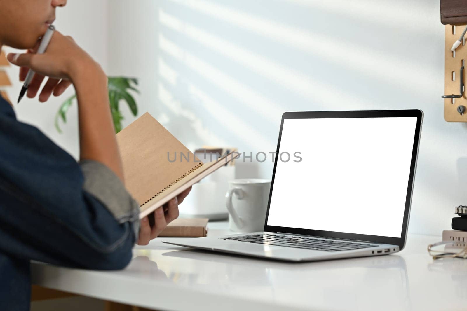 Cropped shot of male freelancer in jeans jacket holding notepad and using laptop on white working desk.
