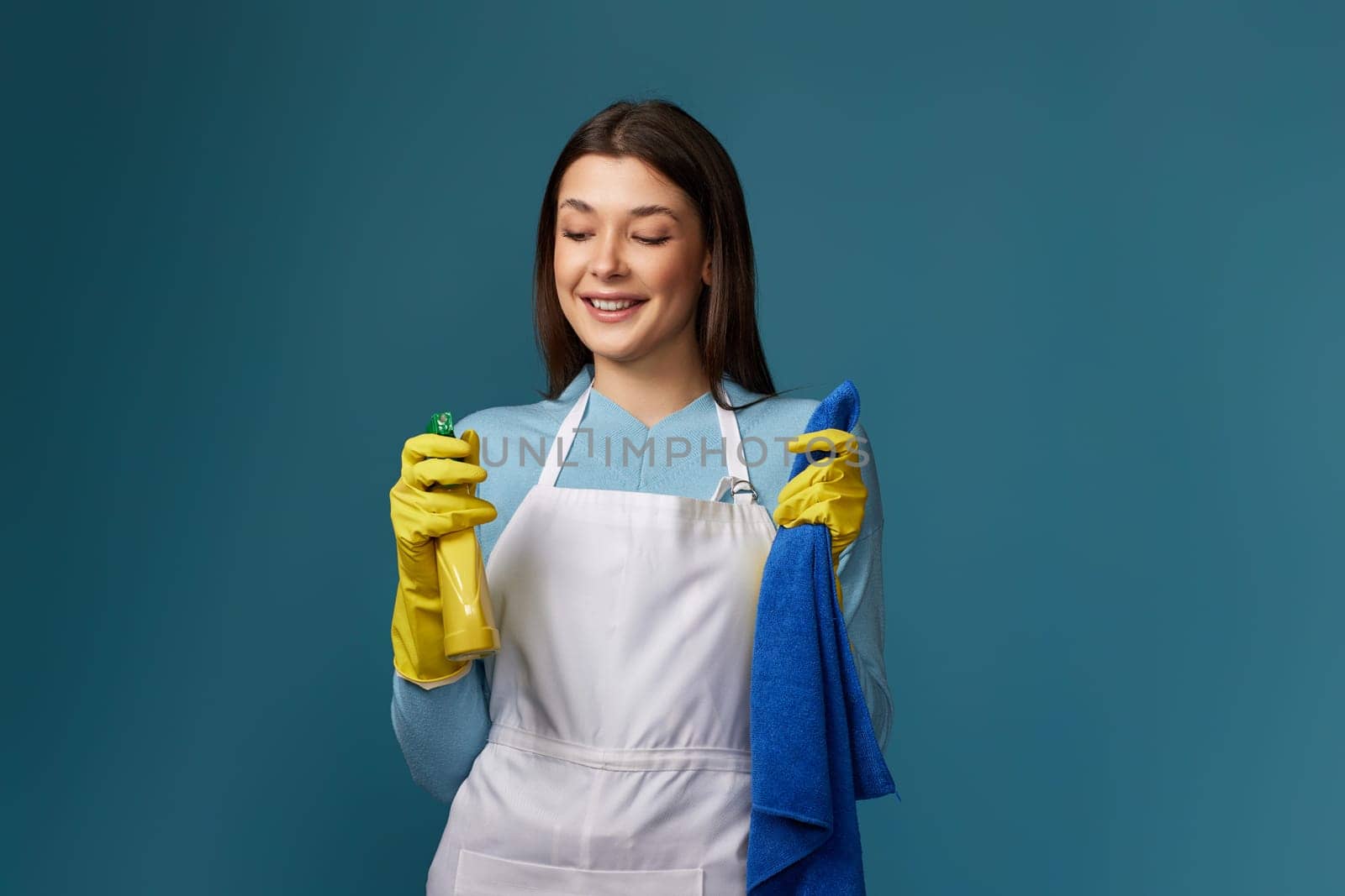 smiling caucasian woman in yellow rubber gloves and cleaner apron with cleaning rag and detergent sprayer on dark blue background.