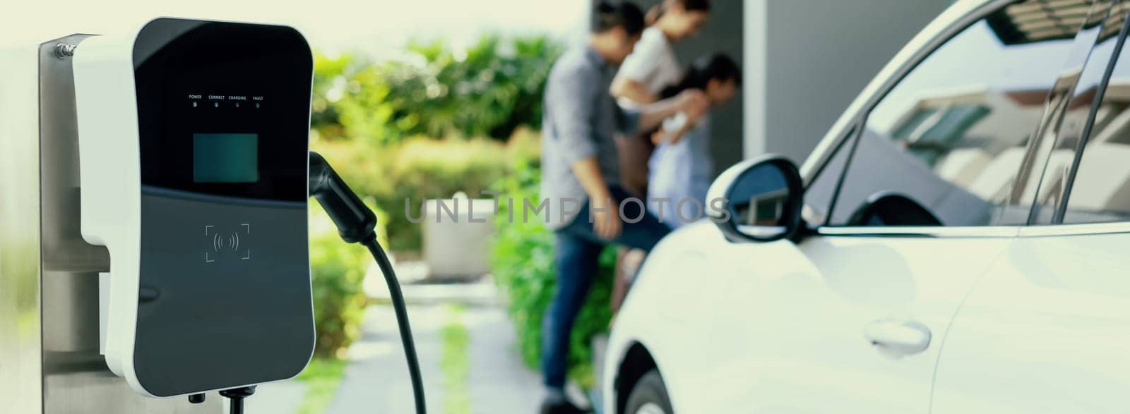 Focus closeup electric vehicle recharging battery from home electric charging station with blurred family in background. Renewable clean energy car for progressive eco awareness lifestyle concept.