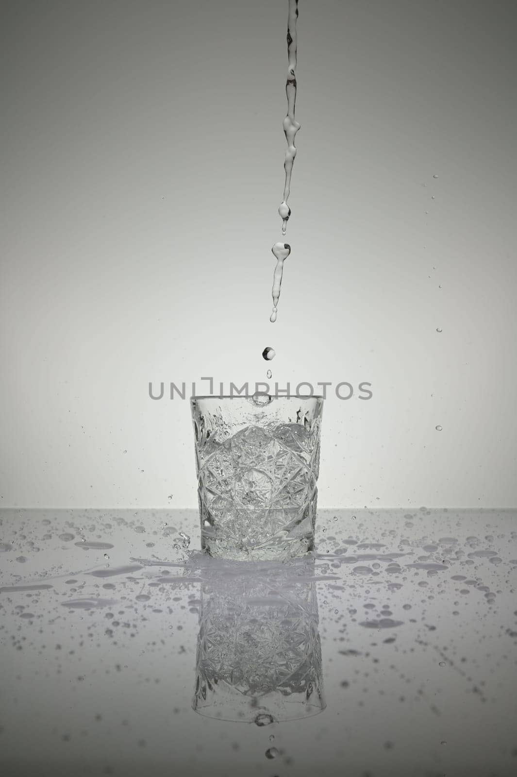 Vertical shot of water pouring into crystal glass with droplets around, on a white shiny table