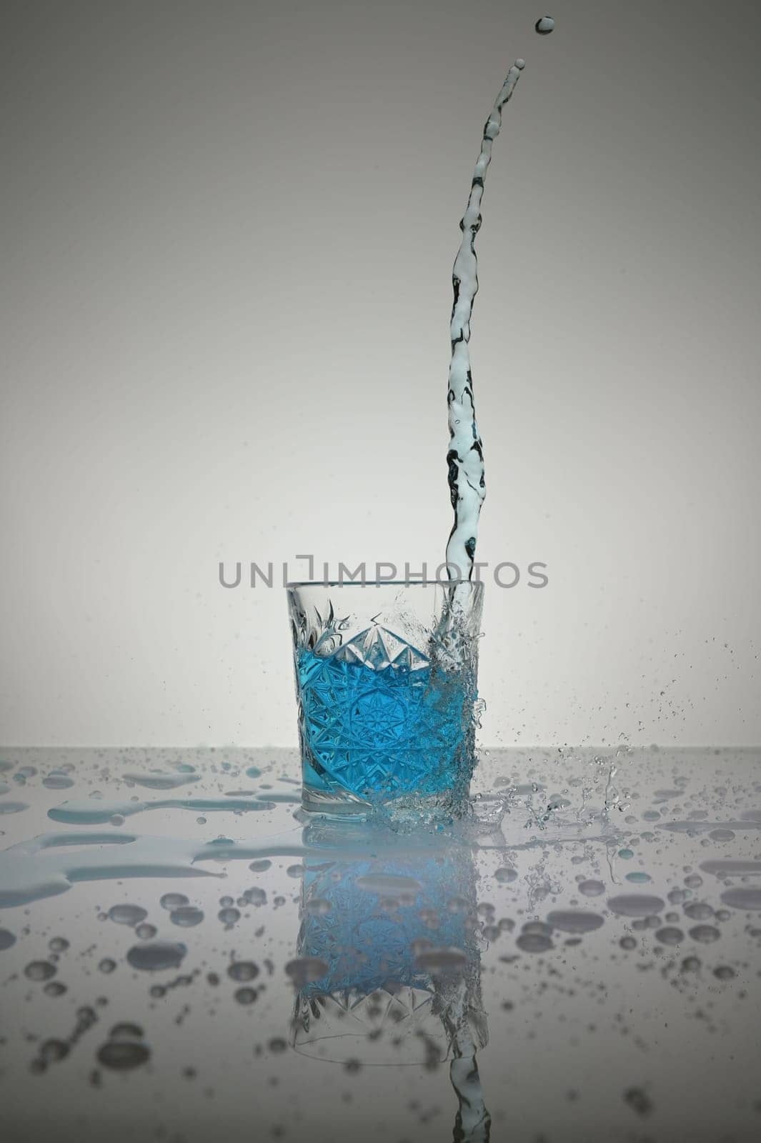 Vertical shot of water pouring into crystal glass with droplets around, on a white shiny table