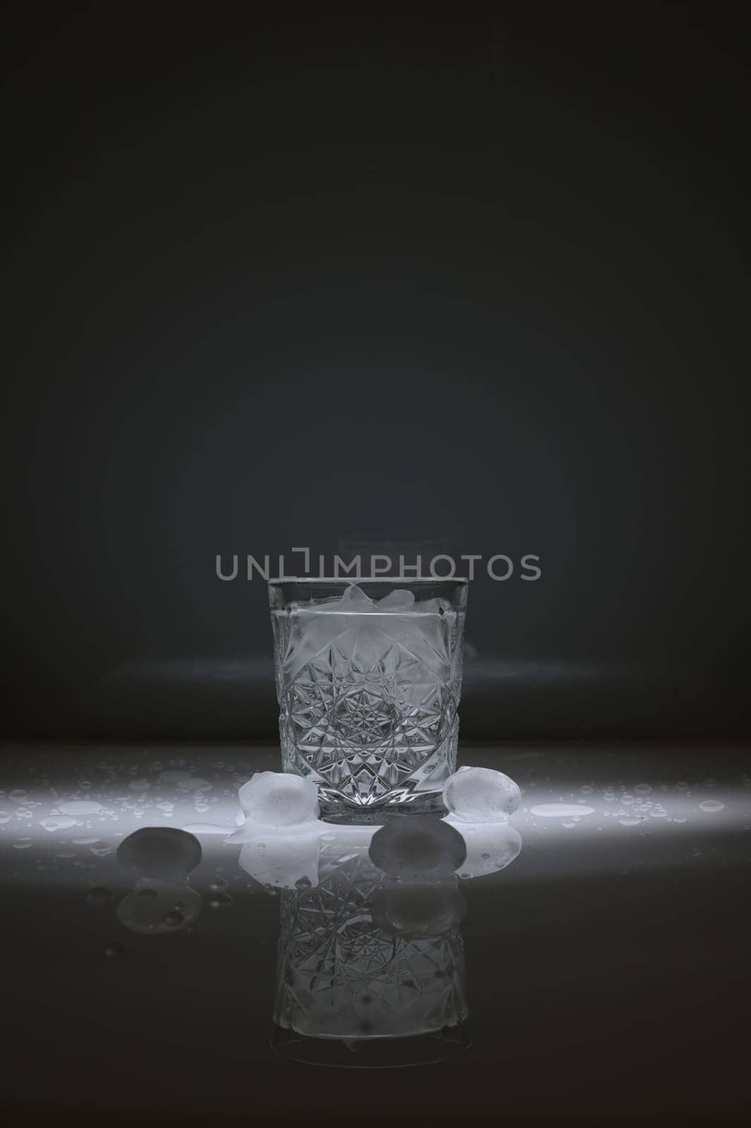 Vertical shot of crystal water glass, ice cubes and waterdrops around, against a dark background