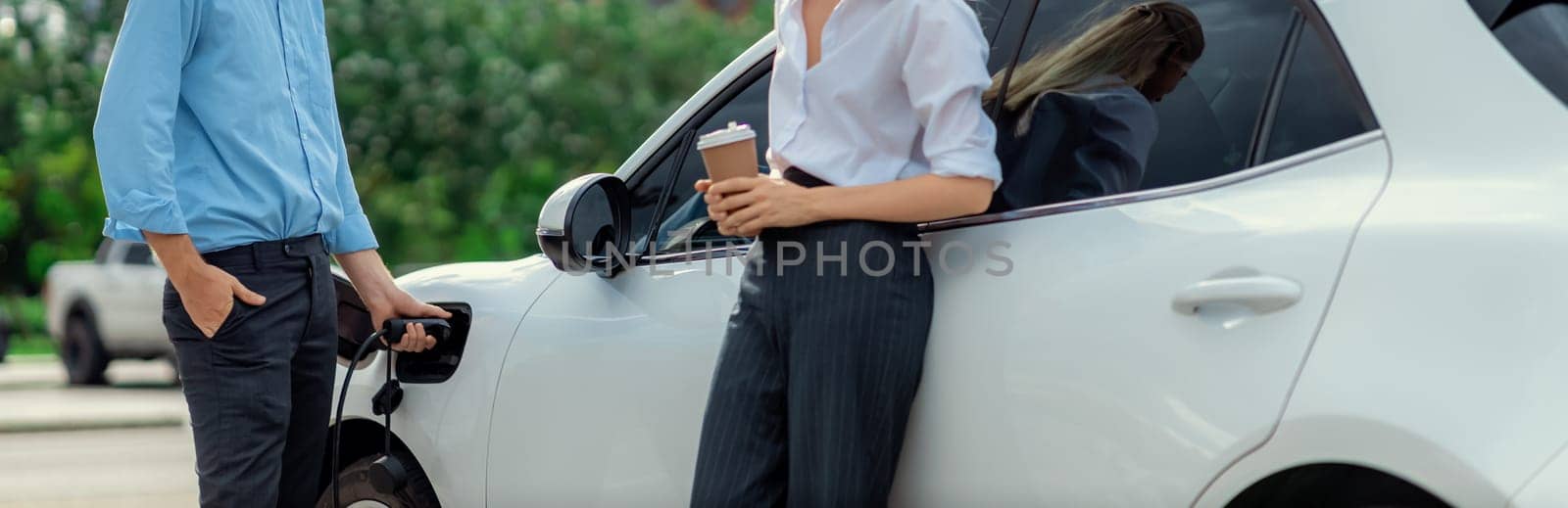 Progressive businessman and businesswoman with coffee, standing at electric car connected to charging station before driving around city center. Eco friendly rechargeable car powered by clean energy.