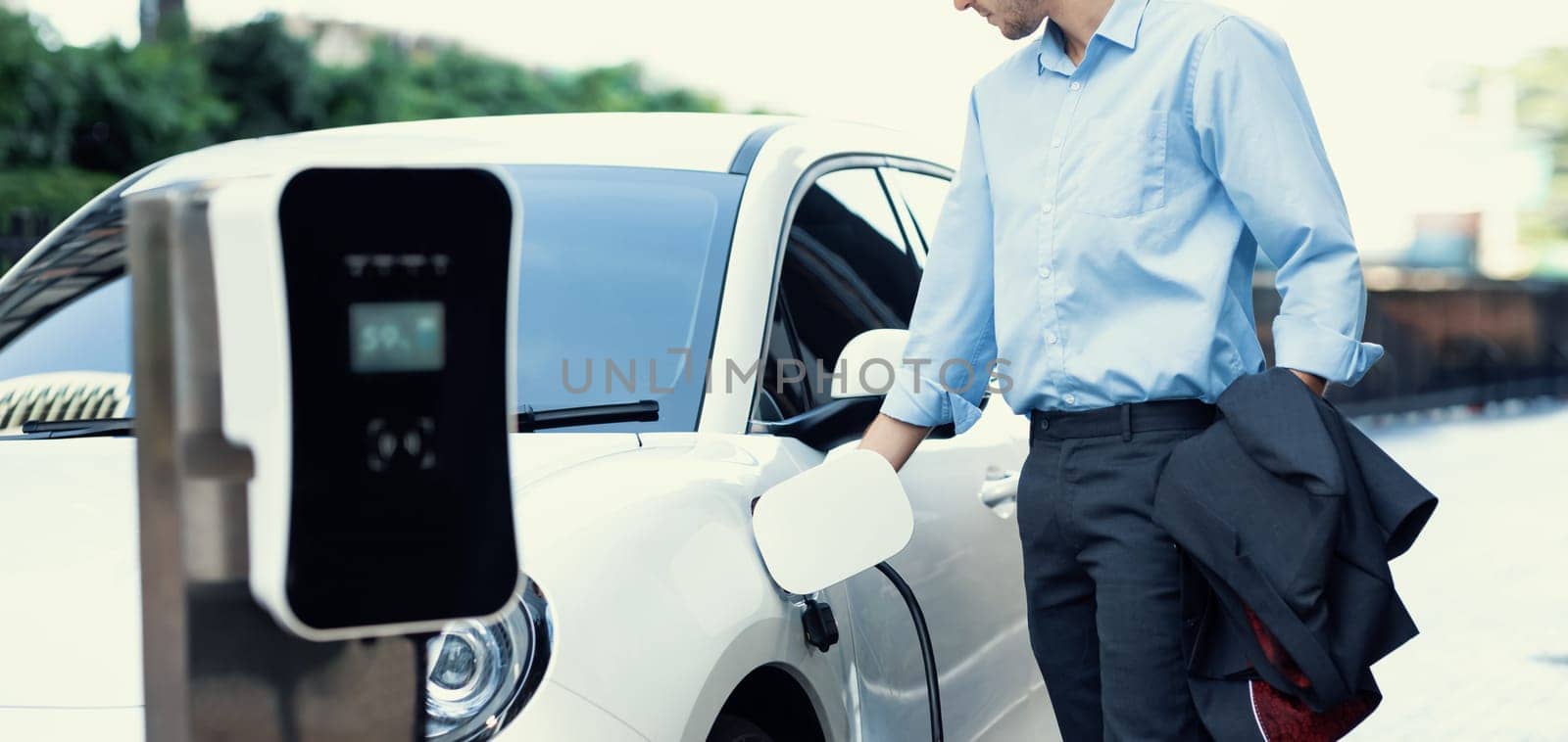 Closeup progressive man holding EV charger plug from public charging station for electric vehicle with background of residential building as concept eco-friendly sustainability energy car concept.