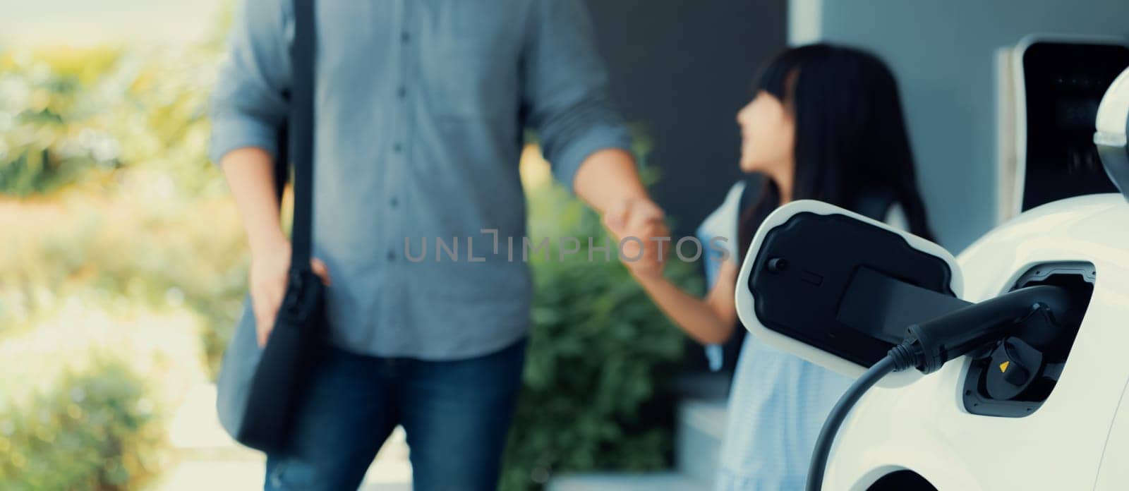 Focus electric car recharging at home charging station with blurred father and daughter walking in background. Progressive green and clean energy vehicle for healthy environment lifestyle concept.