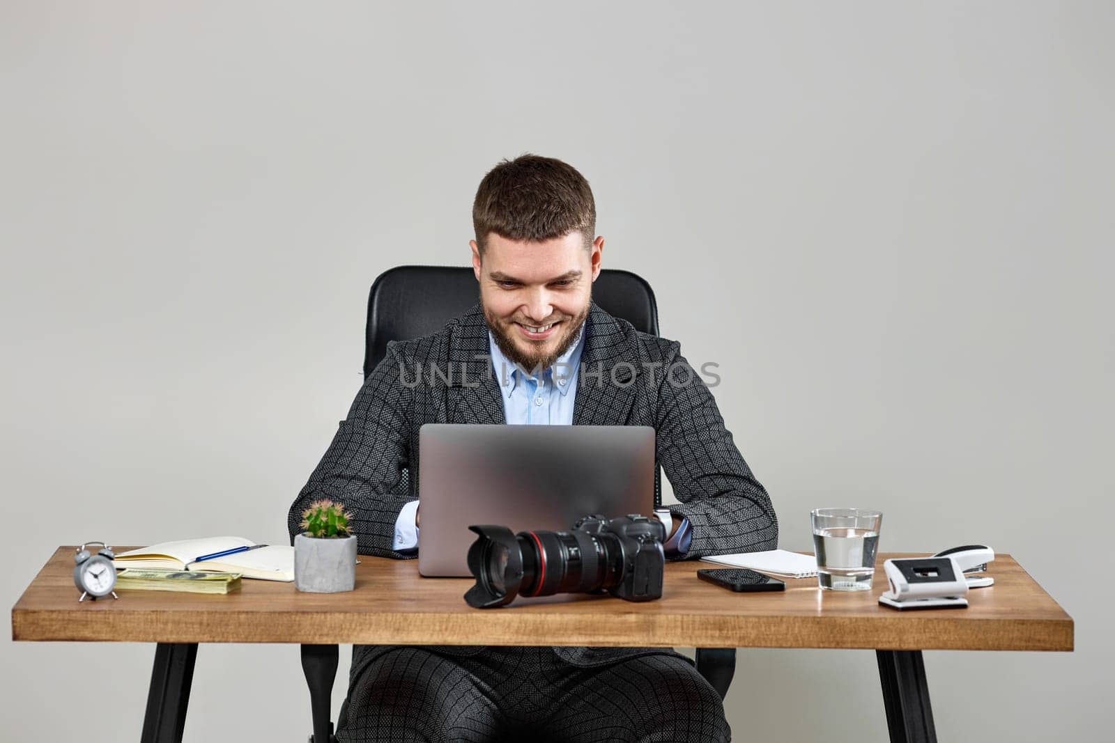 Professional male photographer smiling cheerfully while working on laptop computer at desk. dslr camera on table