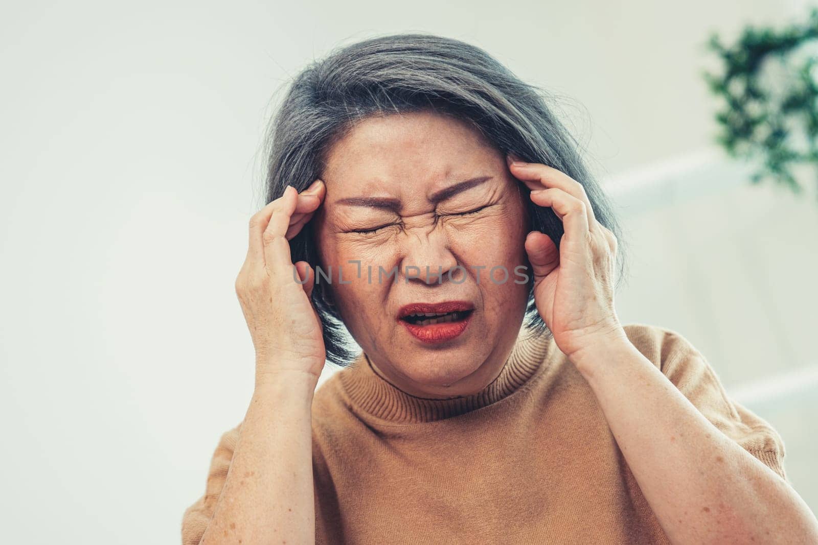 An agonizing senior woman with a headache, compress her temple with both hands at her contented living room. Senior health, nursing home, caretaker service.