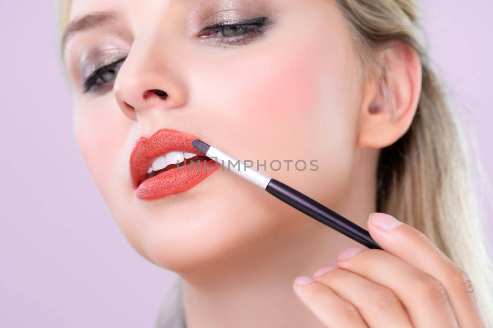 Closeup beautiful young woman with flawless healthy skin and natural makeup putting alluring fashion glossy red lipstick on her lip with lip brush in pink isolated background.