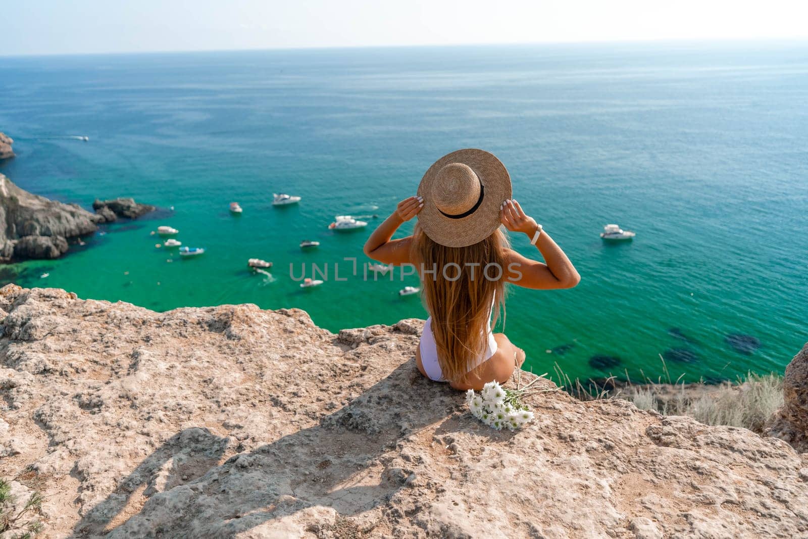 Beach holidays. A hot beautiful woman in a sun hat and a white bikini, sitting with her hands raised to her head, enjoying the view of the ocean on a hot summer day
