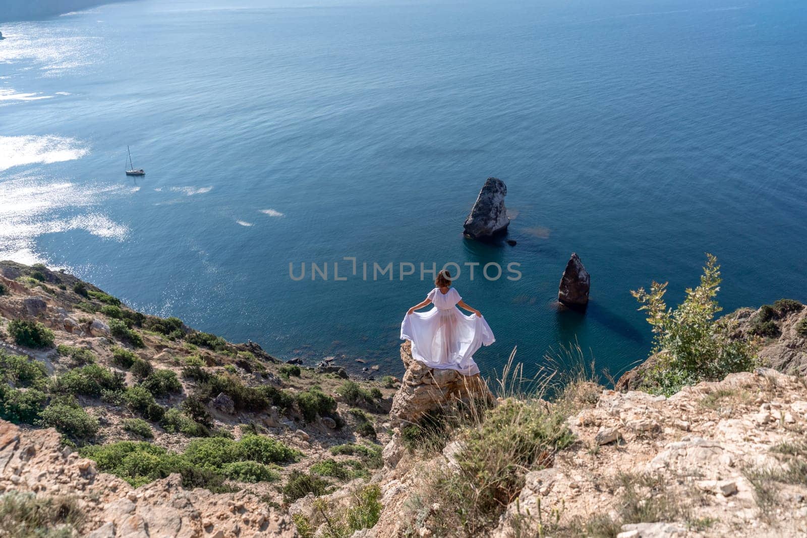 A beautiful young woman in a white light dress with long legs stands on the edge of a cliff above the sea waving a white long dress, against the background of the blue sky and the sea