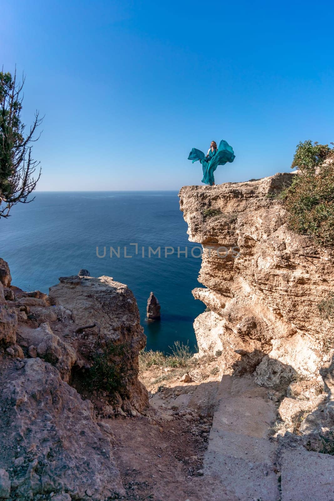 A girl with loose hair in a long mint dress descends the stairs between the yellow rocks overlooking the sea. A rock can be seen in the sea. Sunny path on the sea from the rising sun by Matiunina