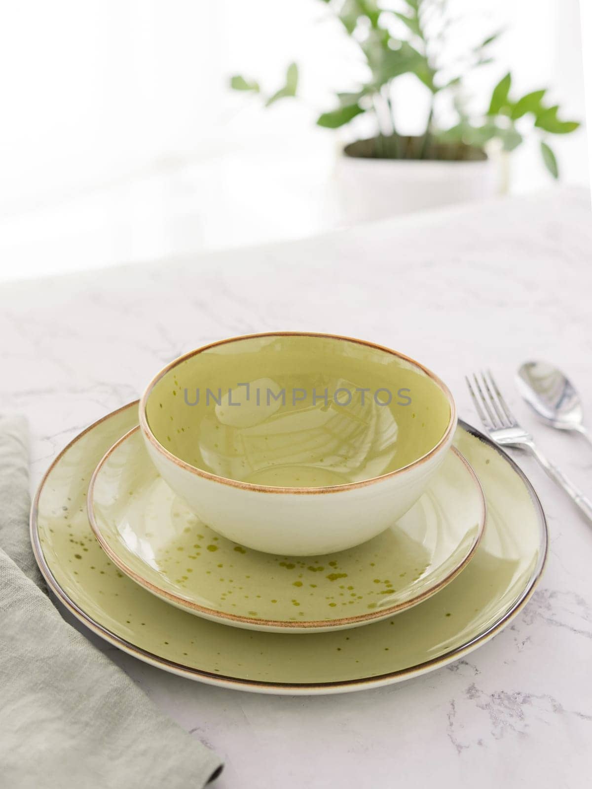 Three empty green plates with silver cutlery and tablecloth on white marble table in front of window by dmitryz