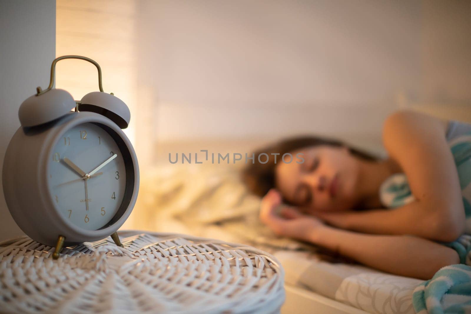 The girl in bed sleeps in the bedroom with an alarm clock on the cabinet next to her. The brunette has her eyes closed and smiles softly in her sleep.