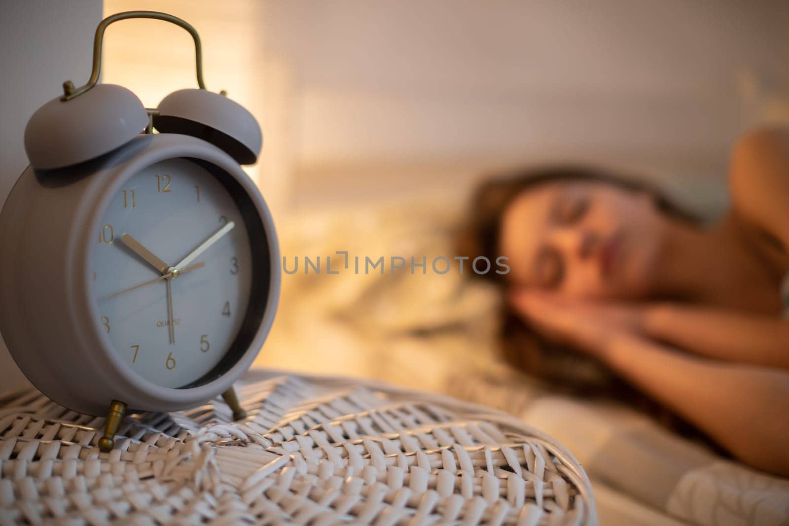 A large alarm clock stands on the cabinet next to the sleeping young woman. A girl under a soft blanket.