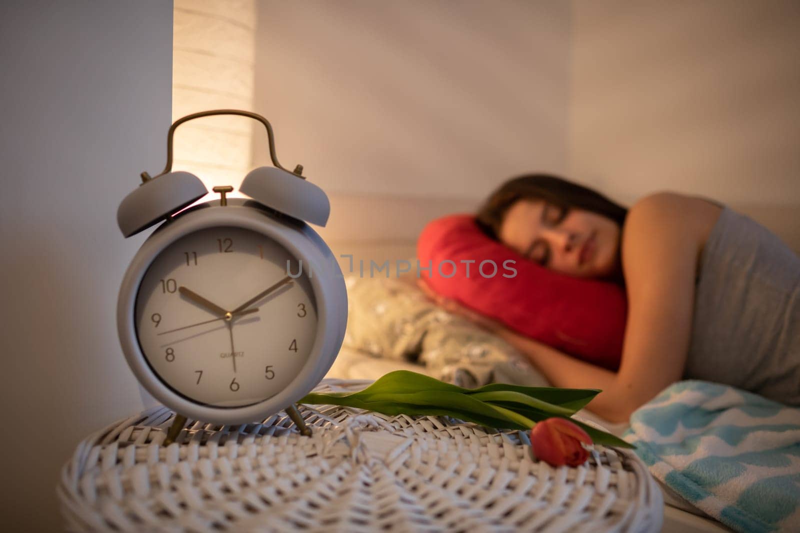 A bedside table with an alarm clock and a tulip stands next to the girl sleeping on her bed. The light of a lamp on the wall.