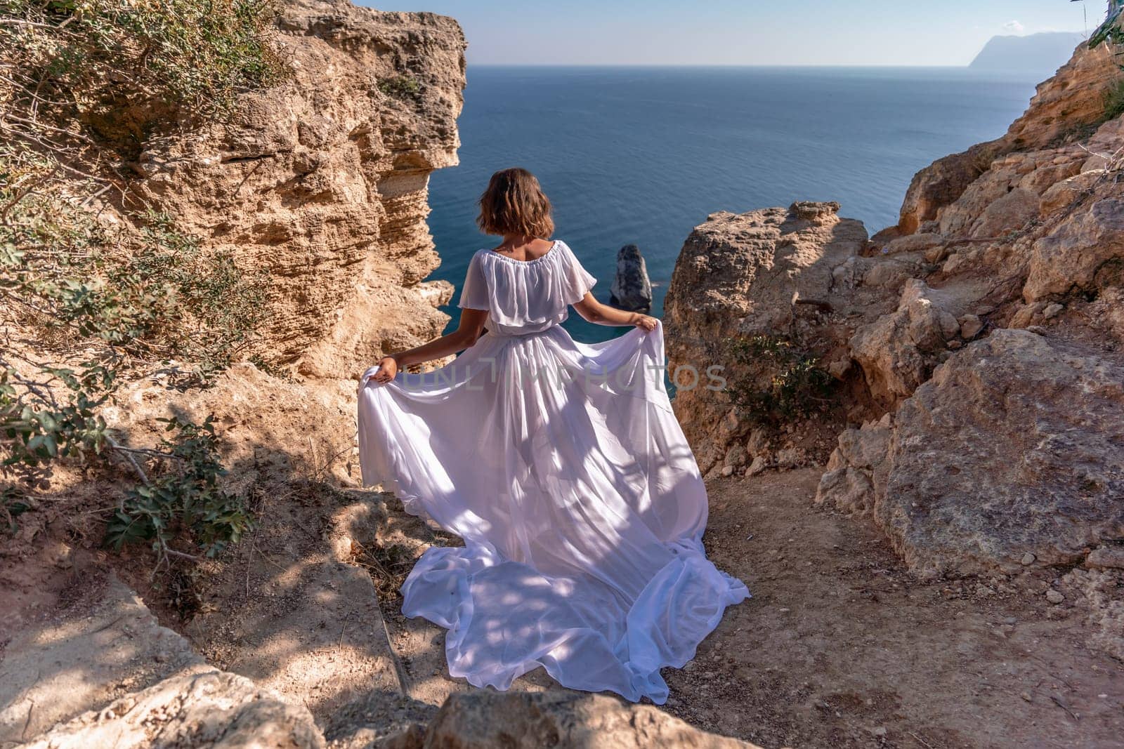 A beautiful young woman in a white light dress with long legs stands on the edge of a cliff above the sea waving a white long dress, against the background of the blue sky and the sea