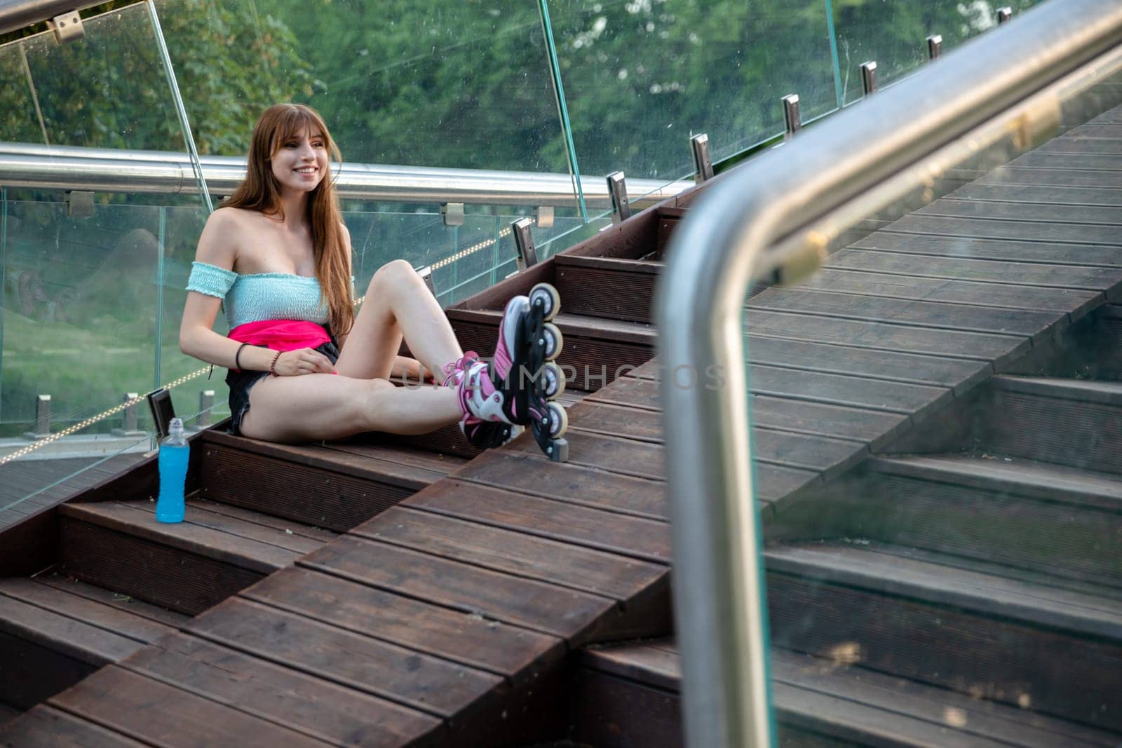 A girl relaxes after rollerblading. A woman sits on a wooden staircase and leans against a glass balustrade. Beside her stands a bottle of blue isotonic drink. In the blurred foreground you can see the railing