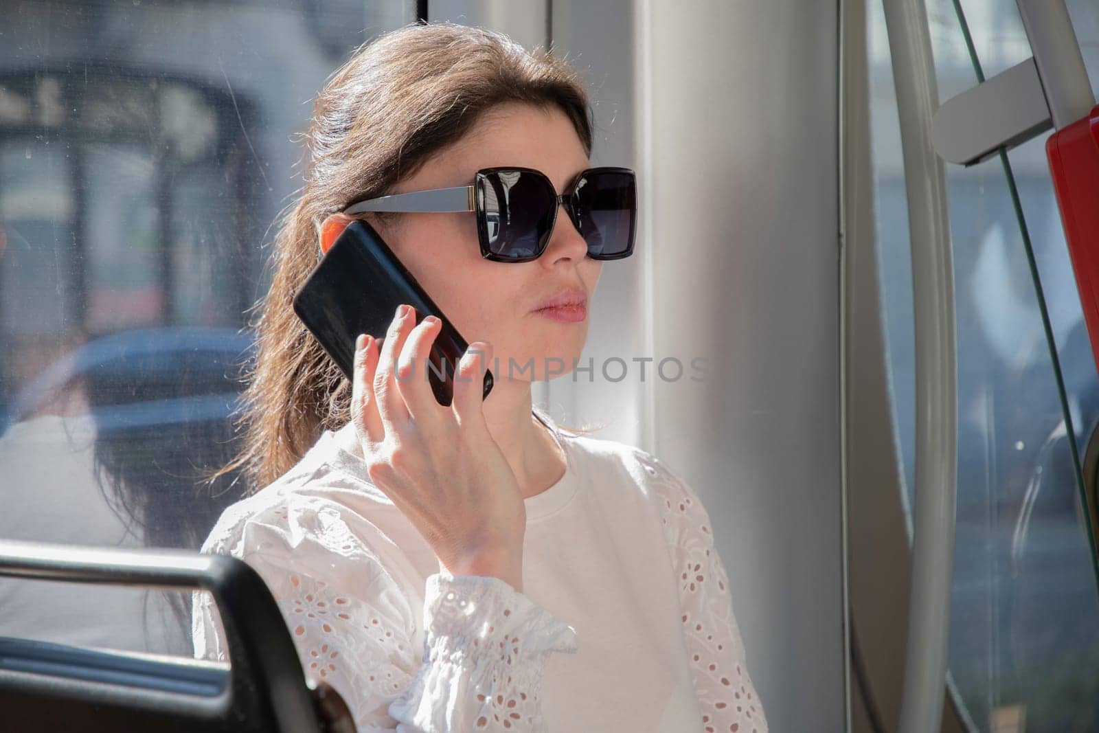 Millennial brunette in sunglasses talking on the phone in a tram by the window, High quality photo