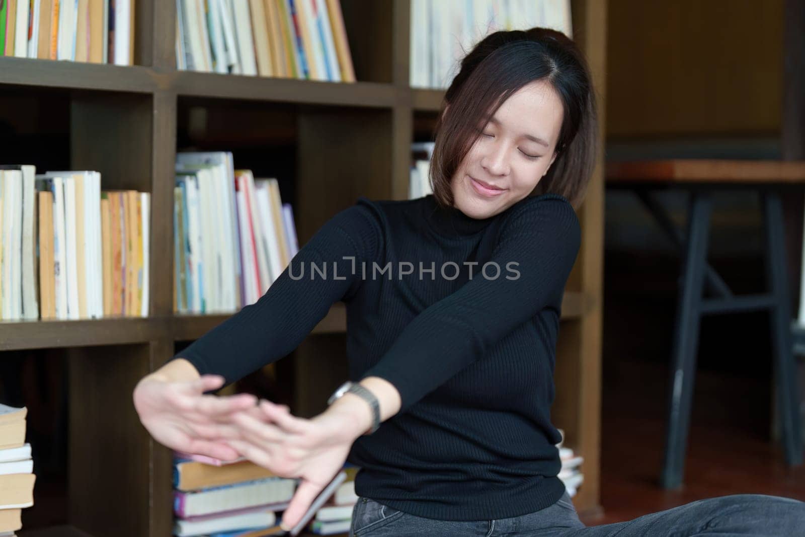 A portrait of a young Asian woman using a computer, wearing headphones and using a notebook to study online shows boredom and pain from video conferencing on a wooden desk in library by Manastrong