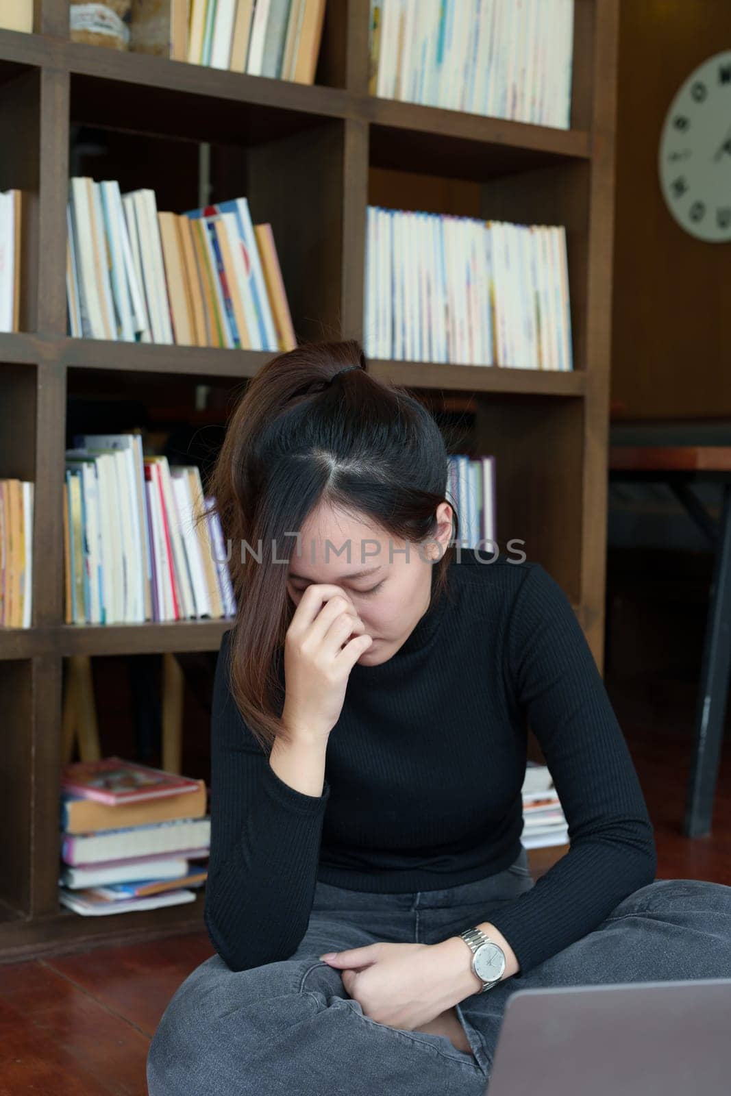 A portrait of a young Asian woman using a computer, wearing headphones and using a notebook to study online shows boredom and pain from video conferencing on a wooden desk in library.