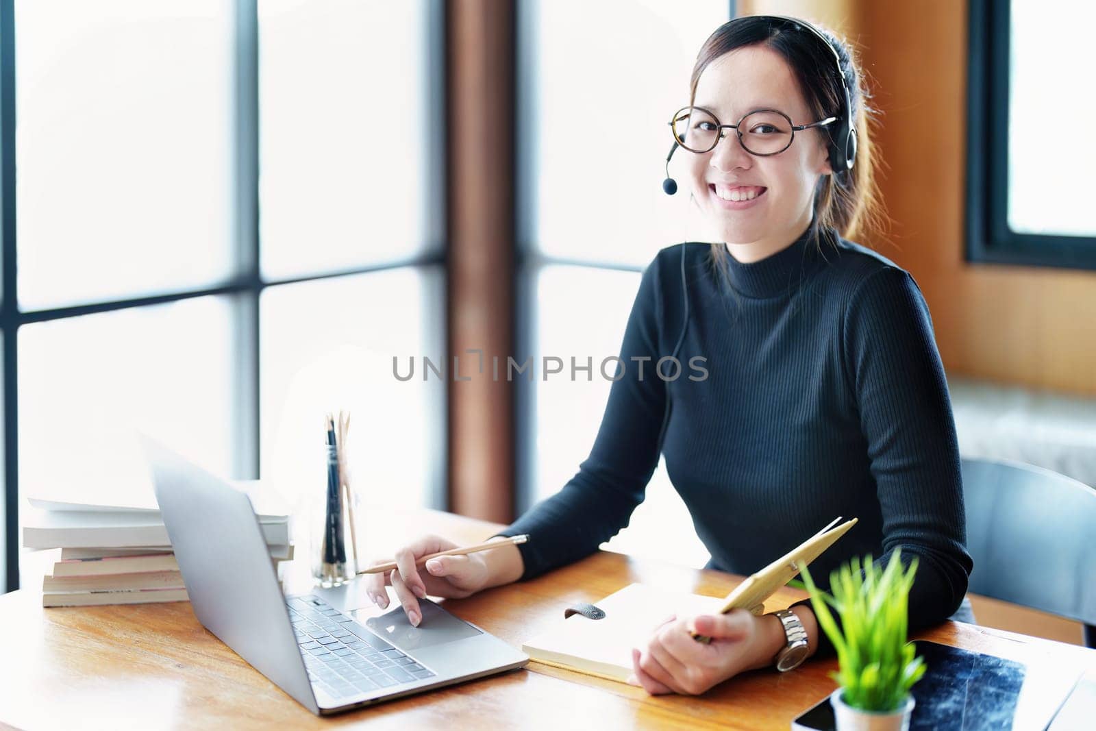 Portrait of a teenage Asian woman wearing glasses using computer laptop, headphones and using a laptop to study online via video conferencing on a wooden library table by Manastrong