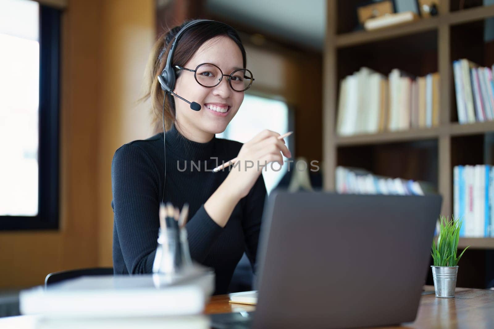 Portrait of a teenage Asian woman wearing glasses using computer laptop, headphones and using a laptop to study online via video conferencing on a wooden library table.