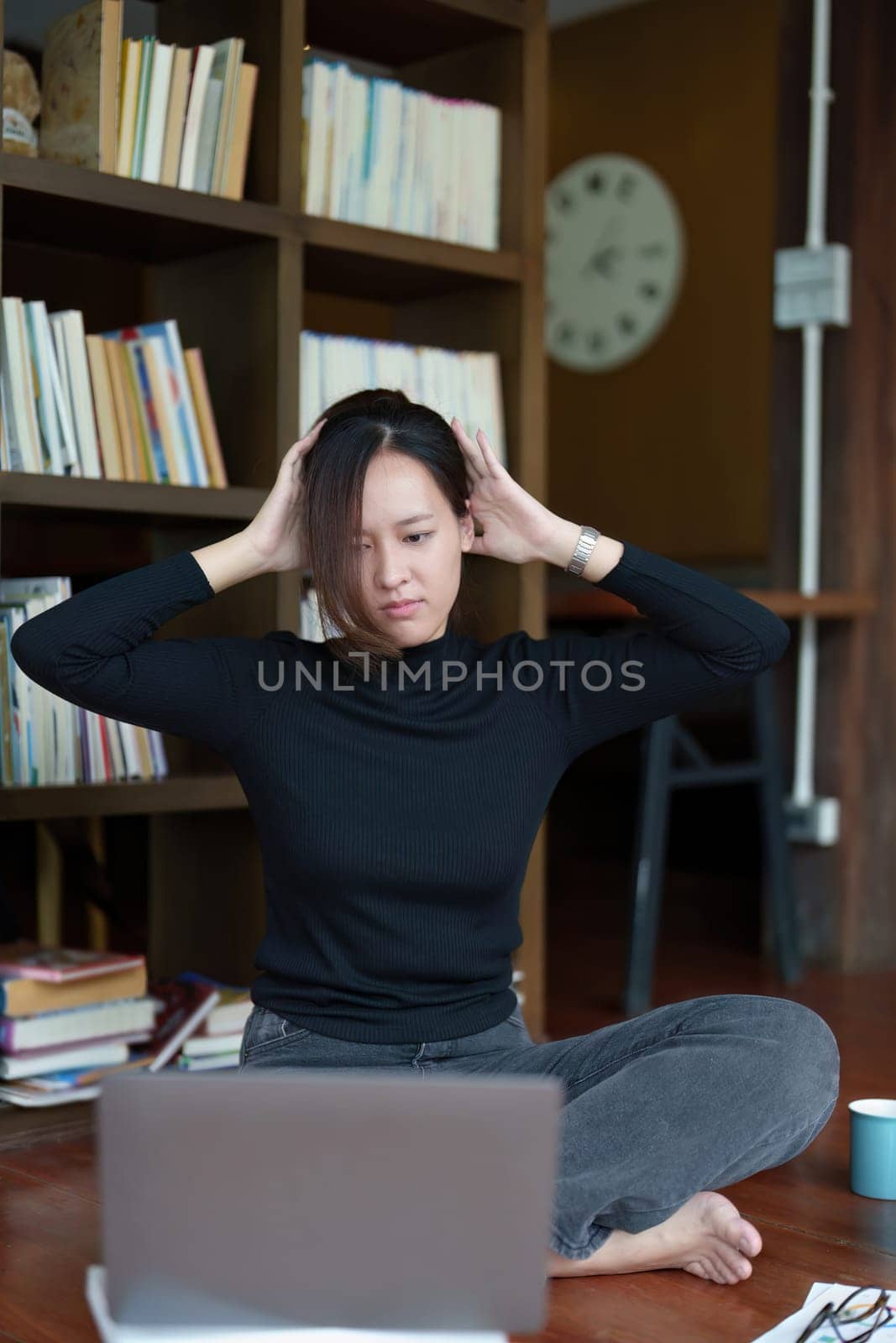 A portrait of a young Asian woman using a computer, wearing headphones and using a notebook to study online shows boredom and pain from video conferencing on a wooden desk in library.