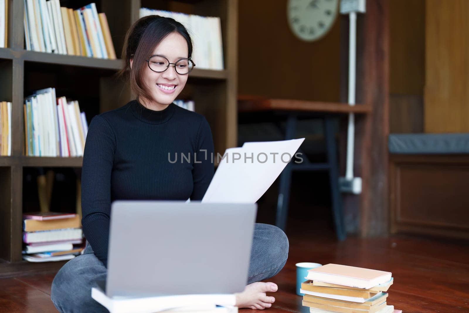 A portrait of a young Asian female student with a smiling face using a computer and holding study materials in the library.