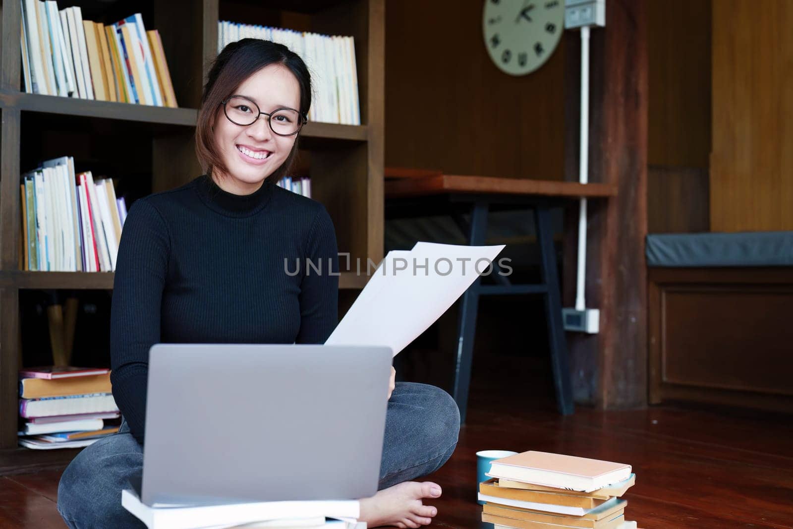 A portrait of a young Asian female student with a smiling face using a computer and holding study materials in the library.