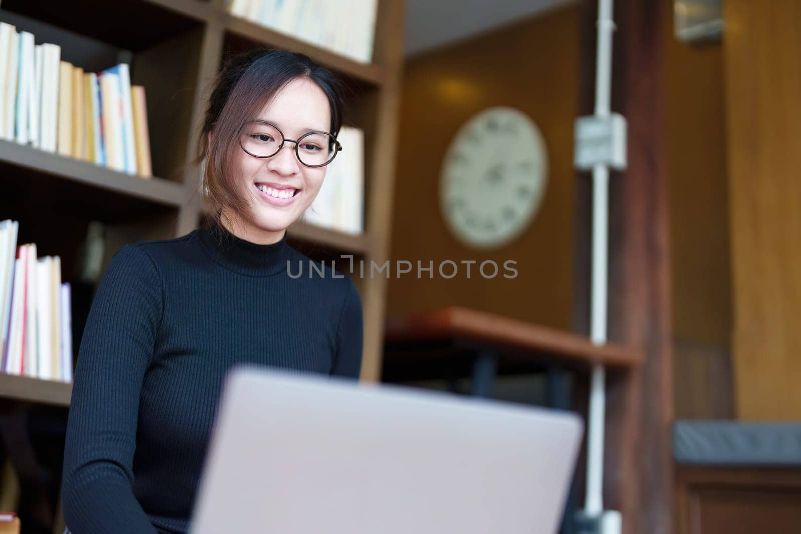 A portrait of a young Asian female student with a smiling face using a computer and holding study materials in the library by Manastrong