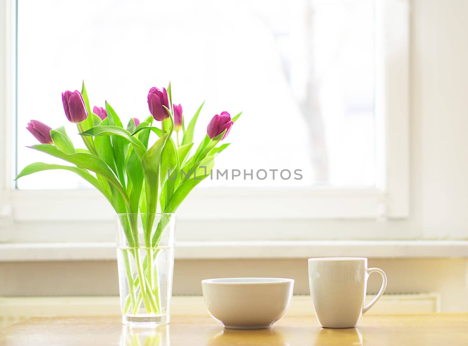 Cozy breakfast with a cup of hot tea or coffee and a bowl of muesli with a magenta tulips bouquet on the wooden table in the front of a window. Good morning. Copy space. High quality photo.