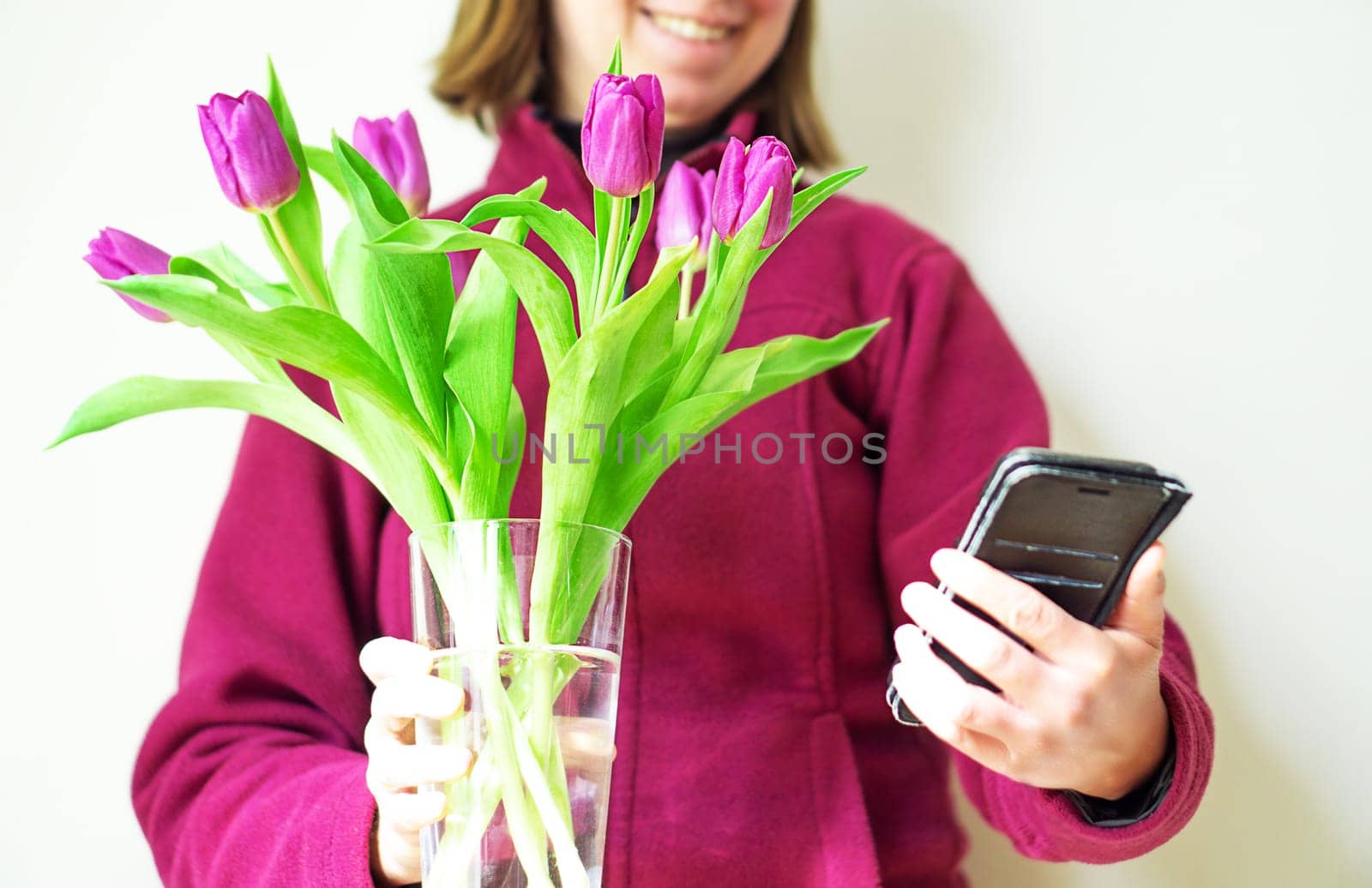 Beautiful girl in cozy purple jacket with magenta tulips is smiling and taking a selfie on her phone by tanjas_photoarts