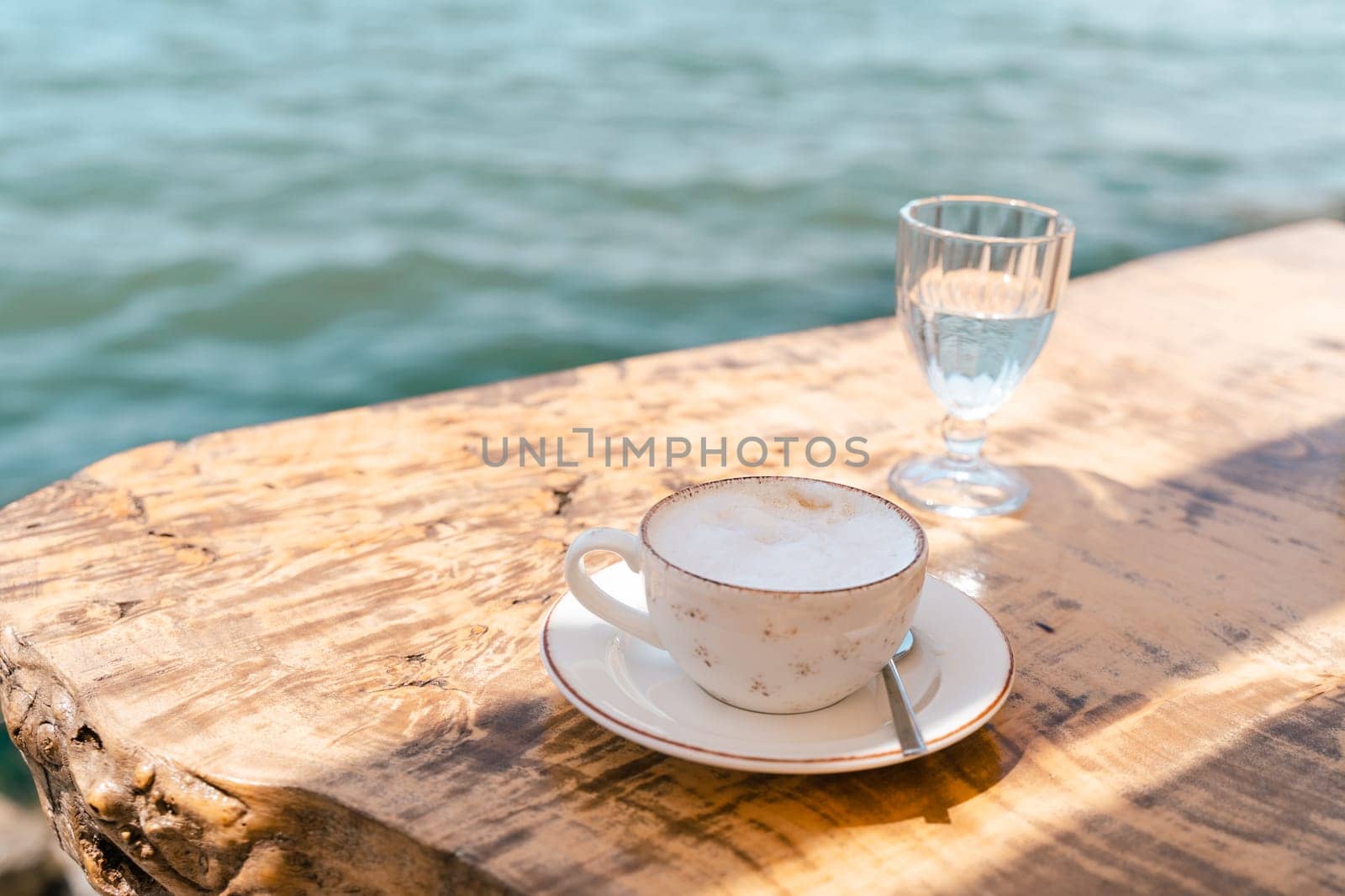 Cappuccino latte coffee cup mug and a glass of water standing on a wooden table in outside cafe with scenery sea and mountains in the background. Copy blank space.