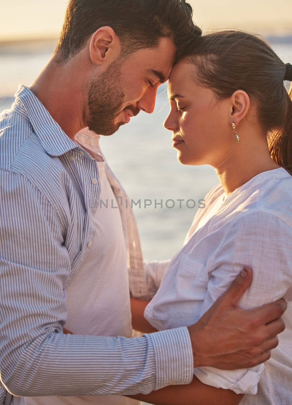 Couple, sunset and beach for love together on holiday or honeymoon. Man, woman and ocean in embrace for romance, during vacation, trip or travel by the sea in late summer sunlight show happiness by YuriArcurs