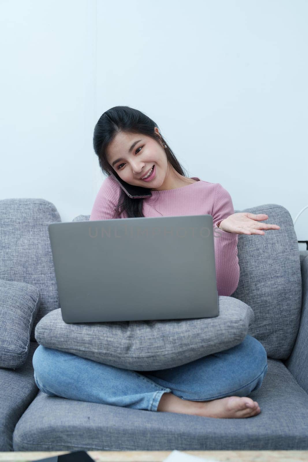 Portrait of a beautiful Asian teenage girl using her phone and computer for video conferencing sitting on the sofa at home.