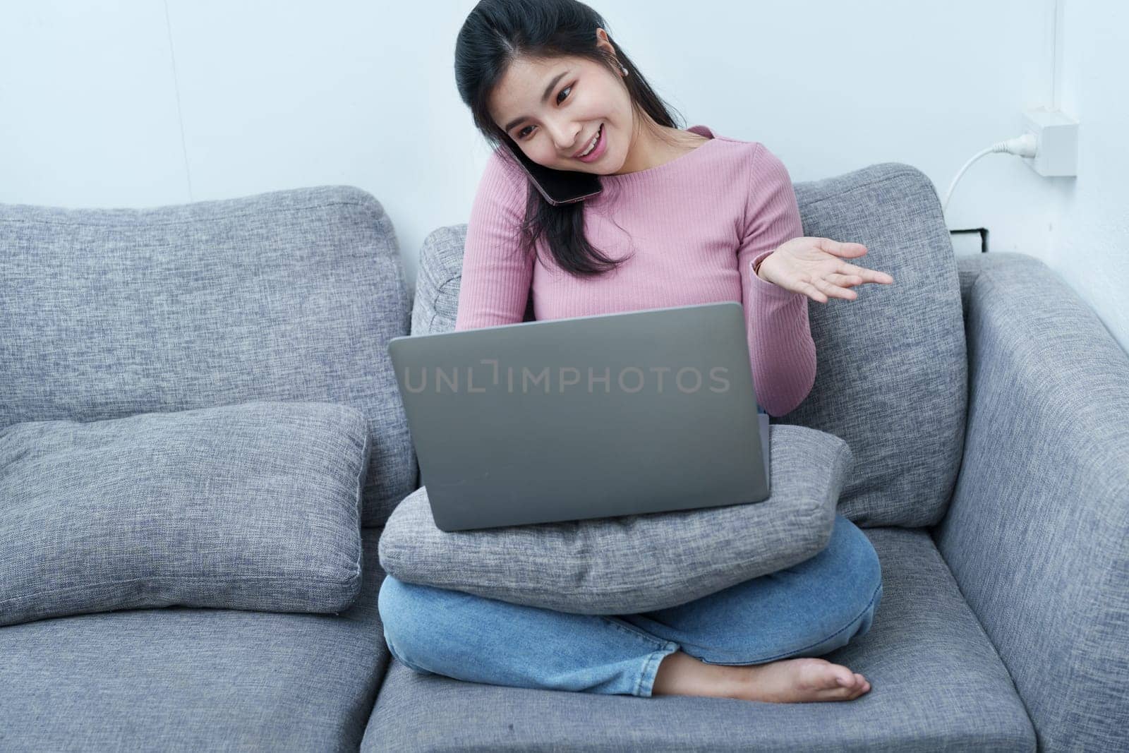 Portrait of a beautiful Asian teenage girl using her phone and computer for video conferencing sitting on the sofa at home by Manastrong