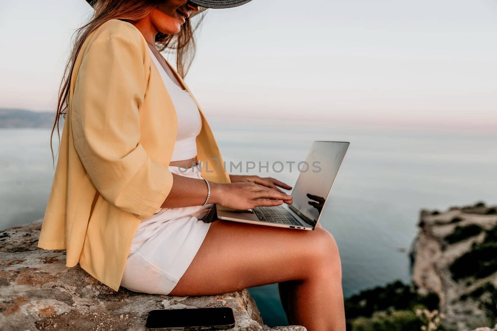 Successful business woman in yellow hat working on laptop by the sea. Pretty lady typing on computer at summer day outdoors. Freelance, travel and holidays concept.