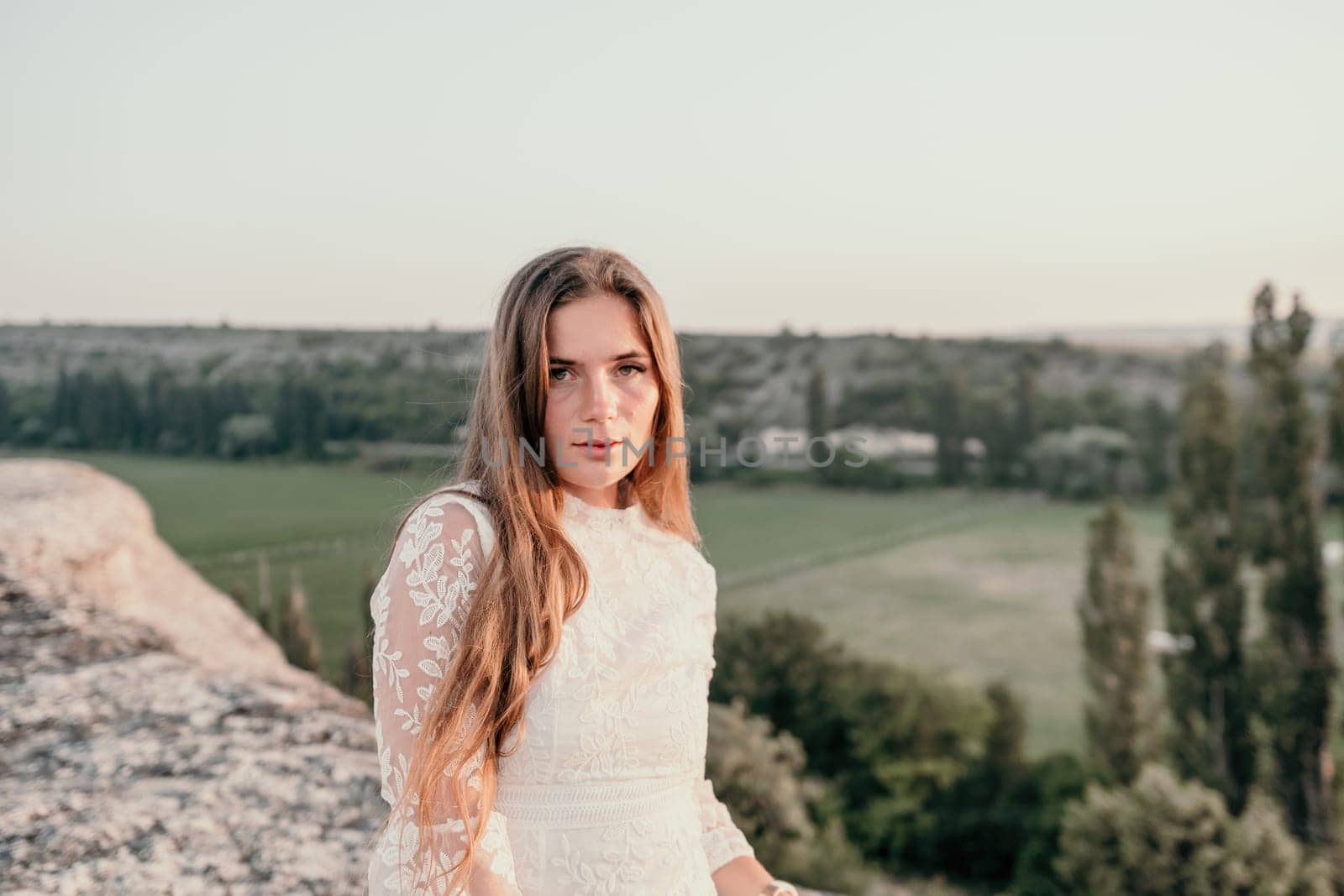 Romantic beautiful bride in white dress posing with sea and mountains in background. Stylish bride standing back on beautiful landscape of sea and mountains on sunset