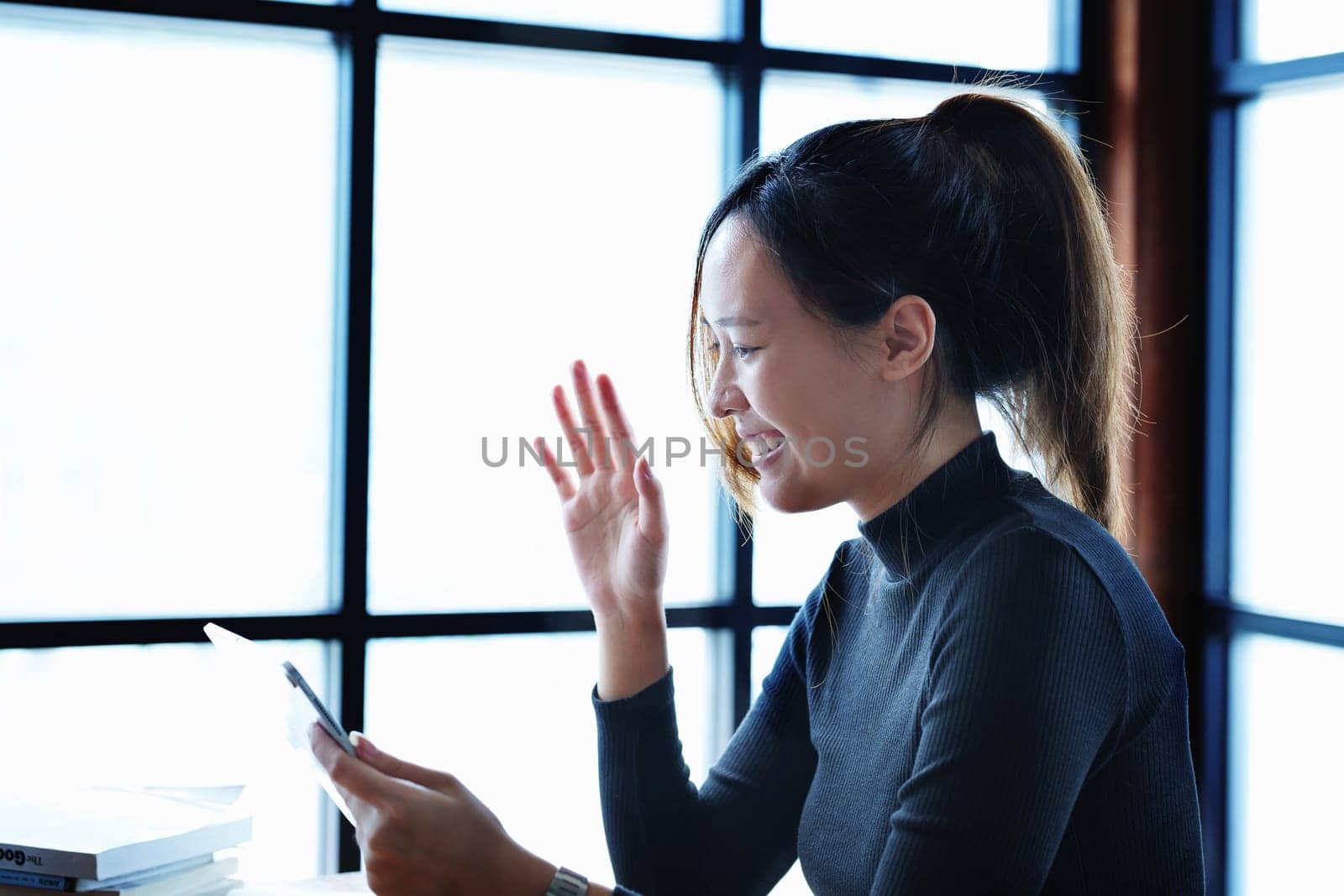 Portrait of a teenage Asian woman using a tablet computer and books to study online via video conferencing on a wooden table in the library.