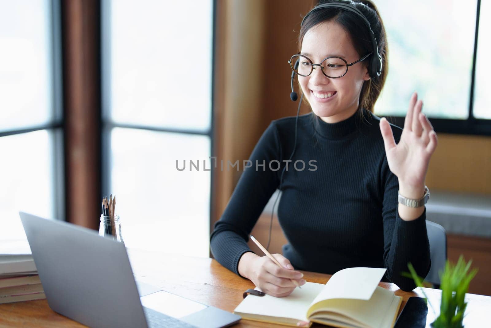 Portrait of a teenage Asian woman wearing glasses using computer laptop, headphones and using a laptop to study online via video conferencing on a wooden library table by Manastrong