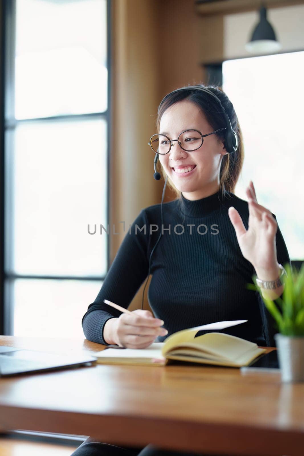 Portrait of a teenage Asian woman wearing glasses using computer laptop, headphones and using a laptop to study online via video conferencing on a wooden library table by Manastrong