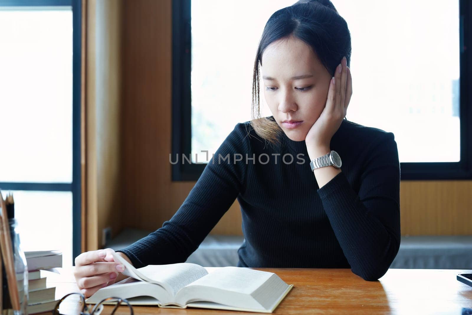 A portrait of a young Asian woman reading textbooks shows boredom on a wooden desk in library..