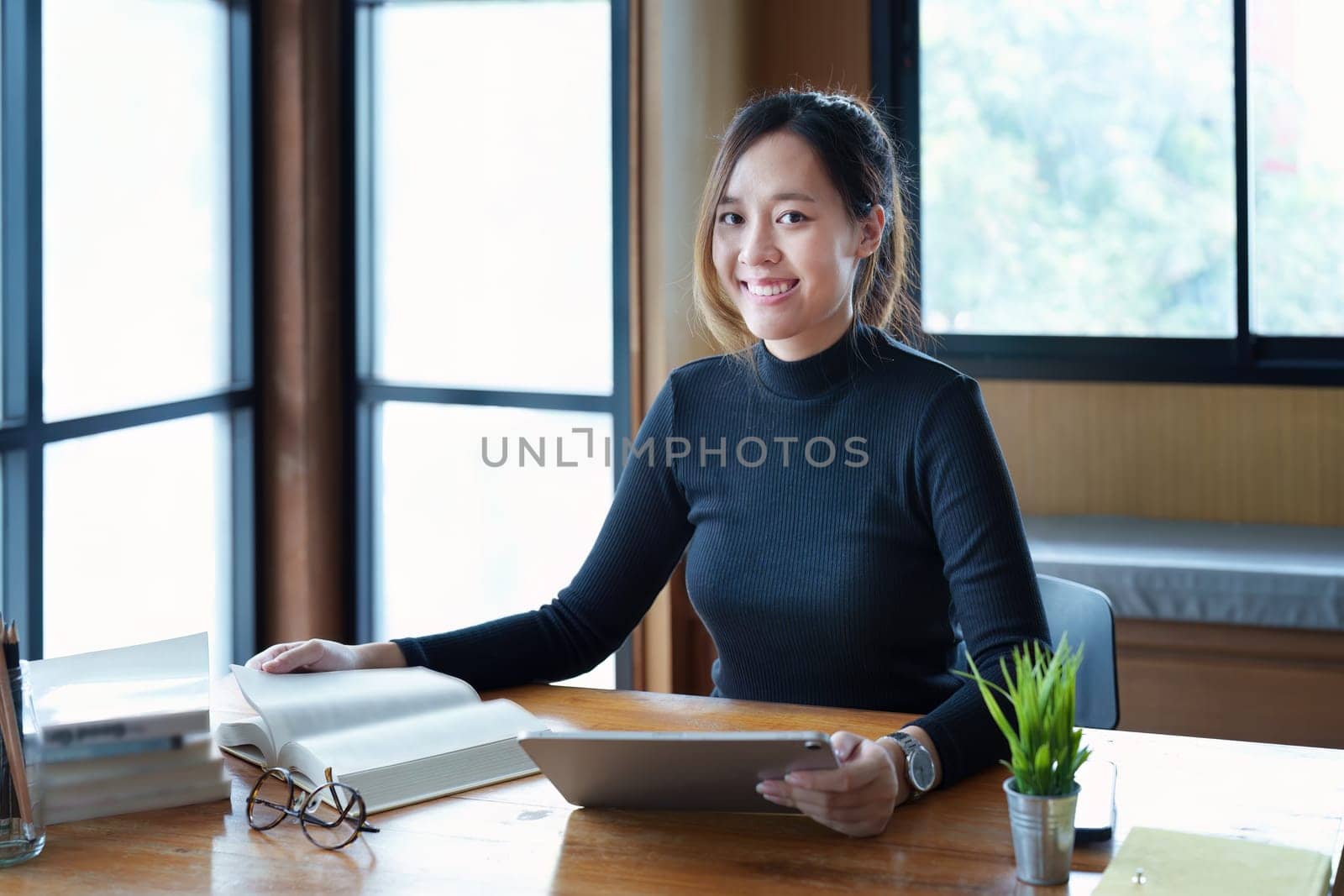 Portrait of a teenage Asian woman using a tablet computer and books to study online via video conferencing on a wooden table in the library.