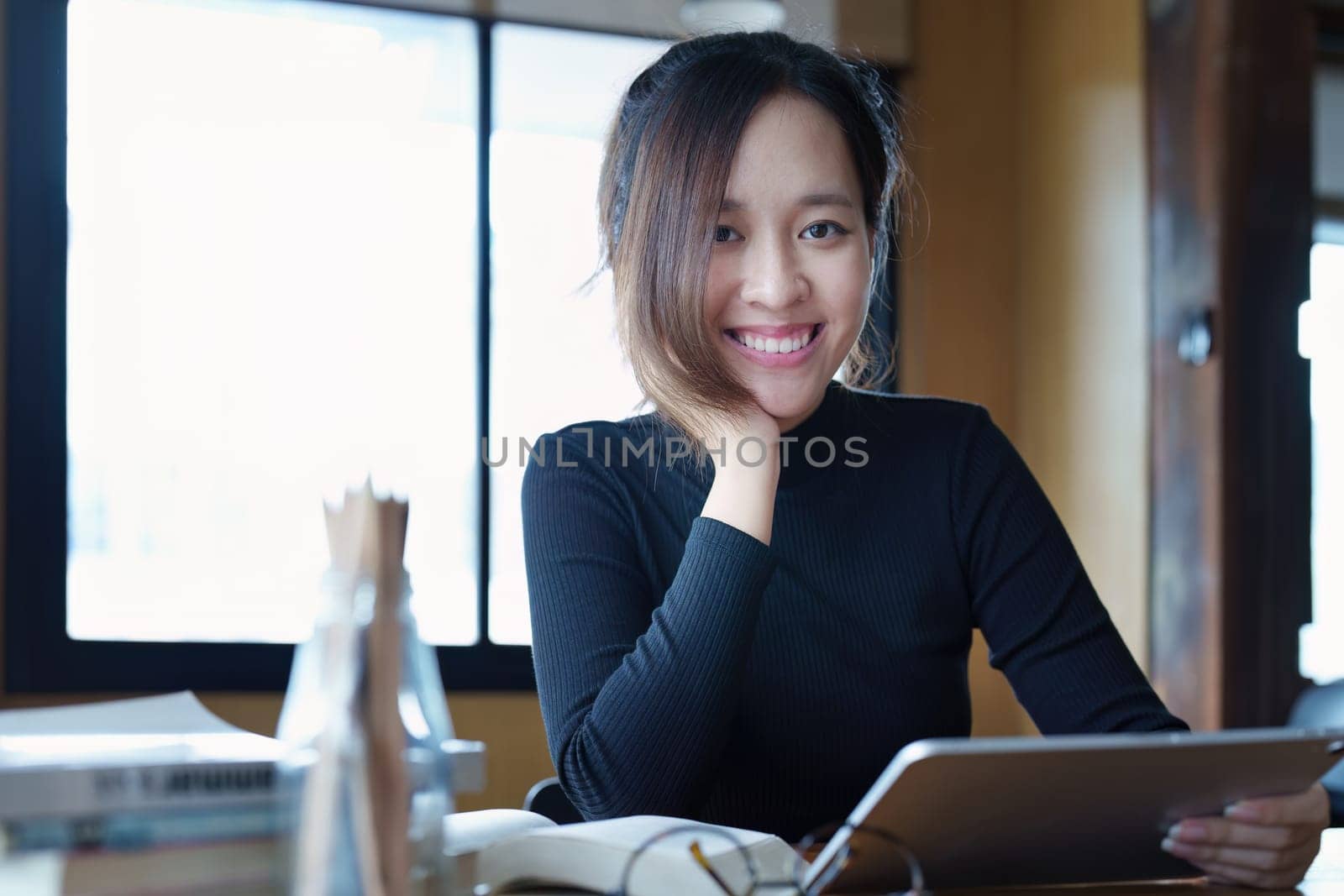Portrait of a teenage Asian woman using a tablet computer and books to study online via video conferencing on a wooden table in the library.