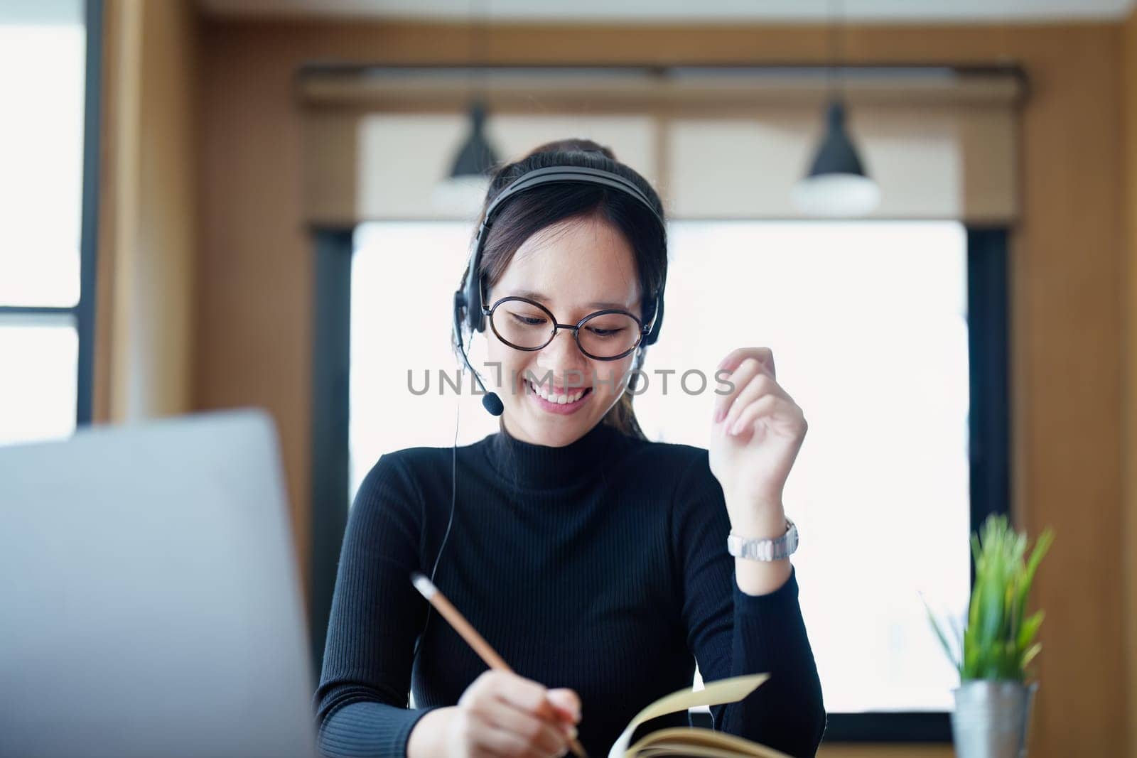 Portrait of a teenage Asian woman wearing glasses using computer laptop, headphones and using a laptop to study online via video conferencing on a wooden library table.