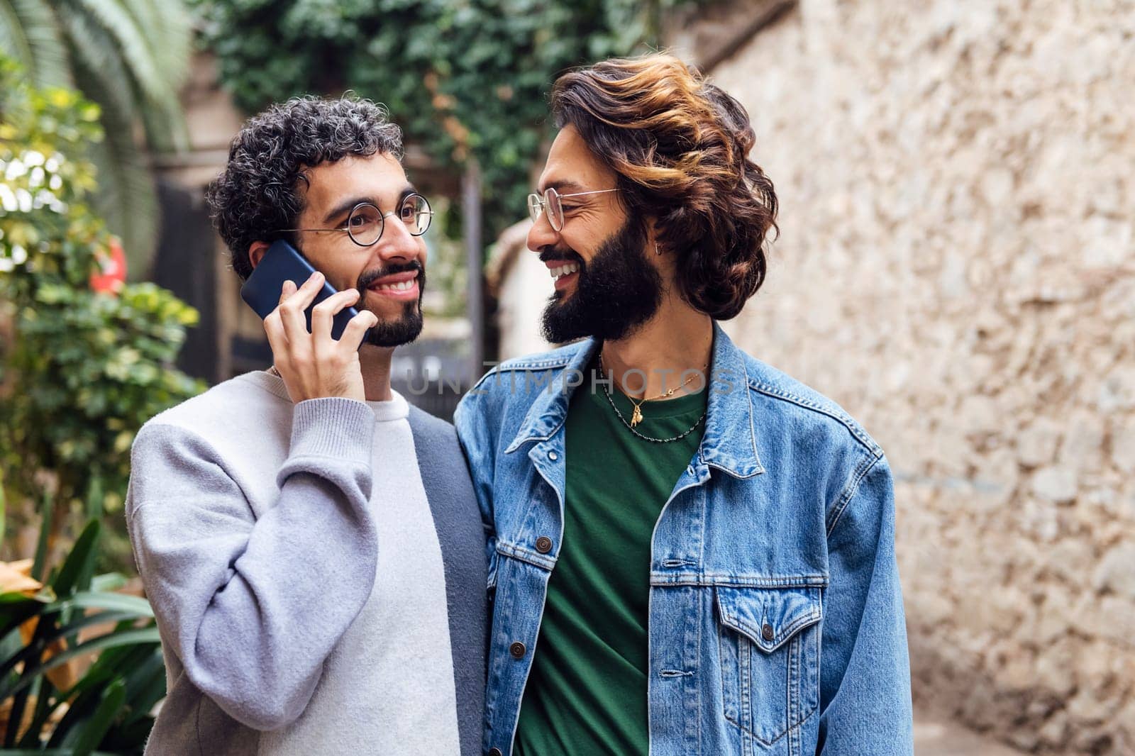 gay male couple smiling happily while one of them is talking on the phone in a nice street with plants, concept of communication and love between people of the same sex