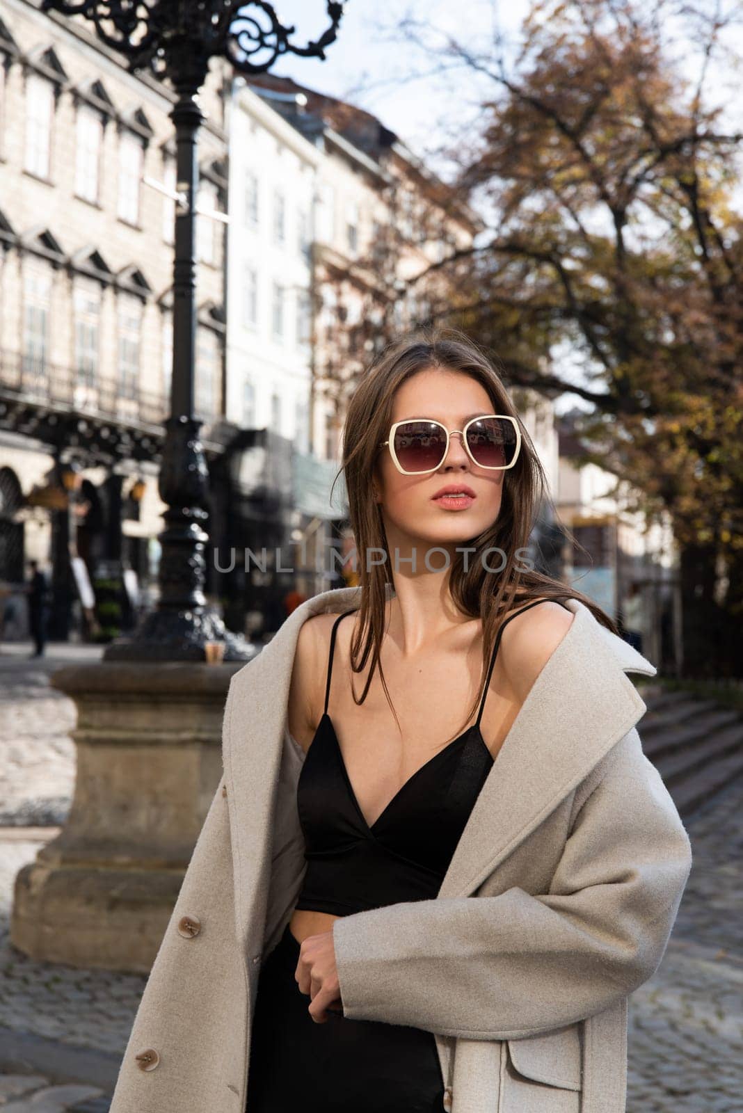 charming brunette posing on the street in spring, wearing a stylish beige coat, a top with razors and sunglasses by Ashtray25