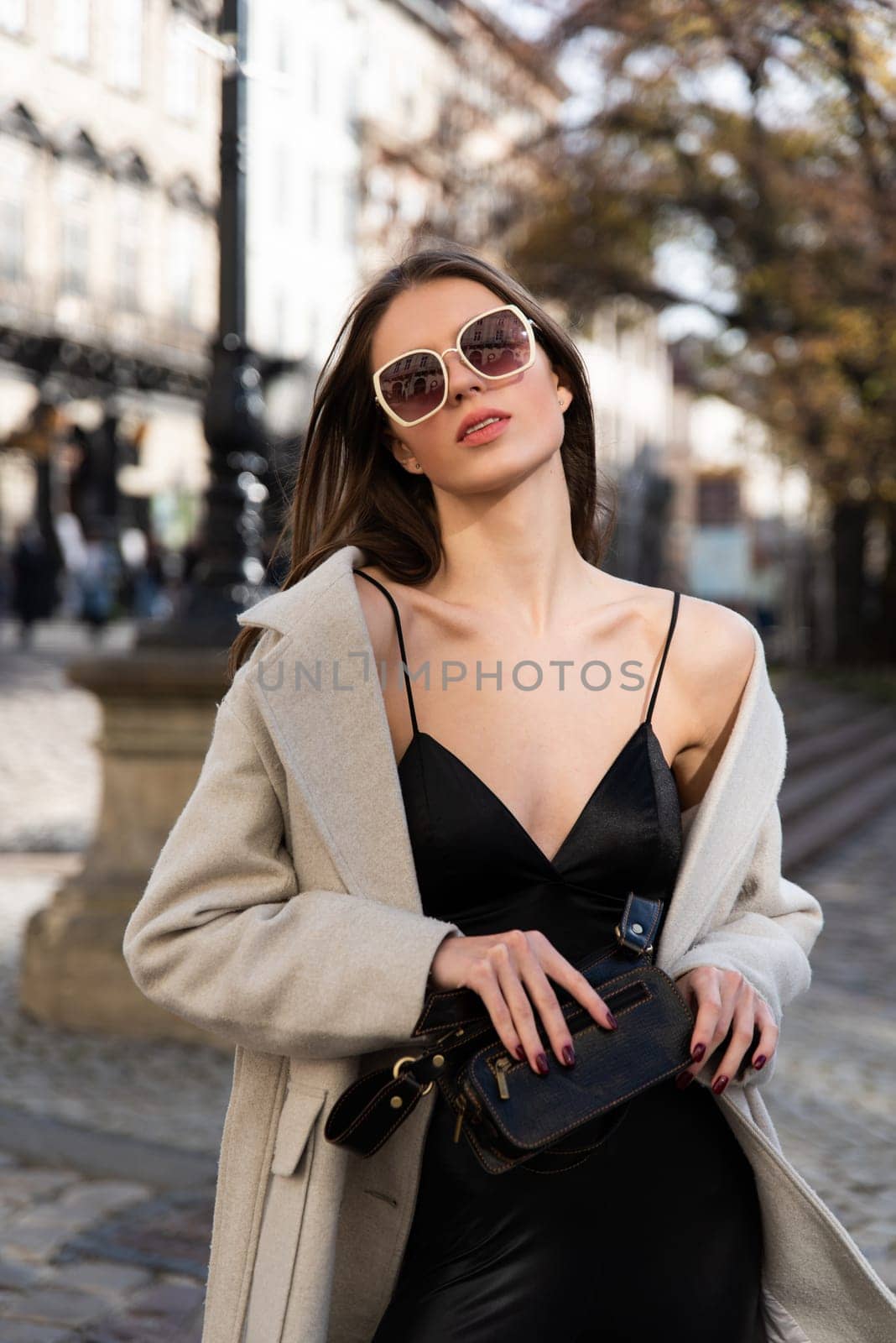 charming brunette posing on the street in spring, wearing a stylish beige coat, a top with razors and sunglasses